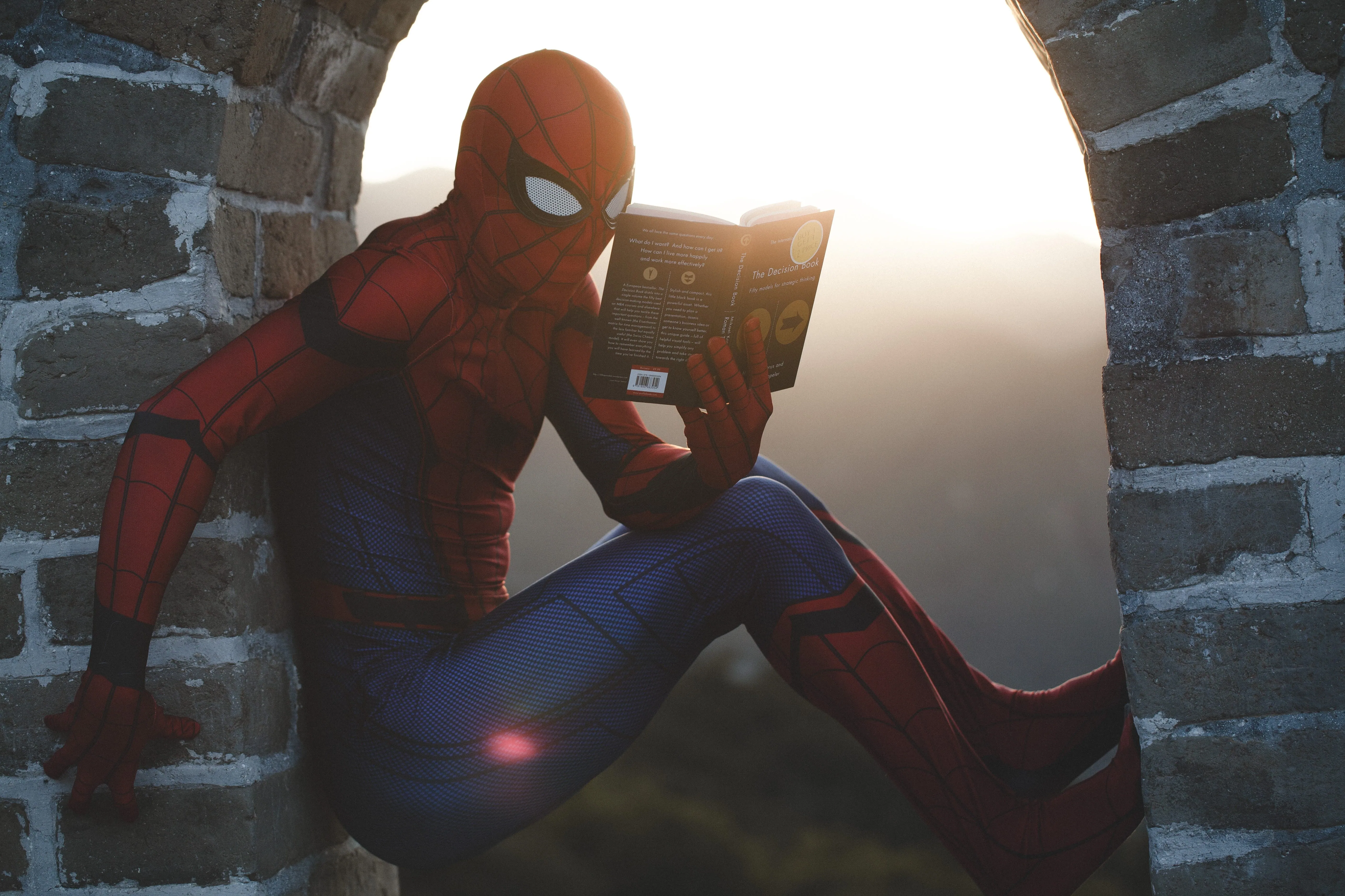 A person in a Spider-Man costume perched in a brick archway at sunset, reading The Decision Book