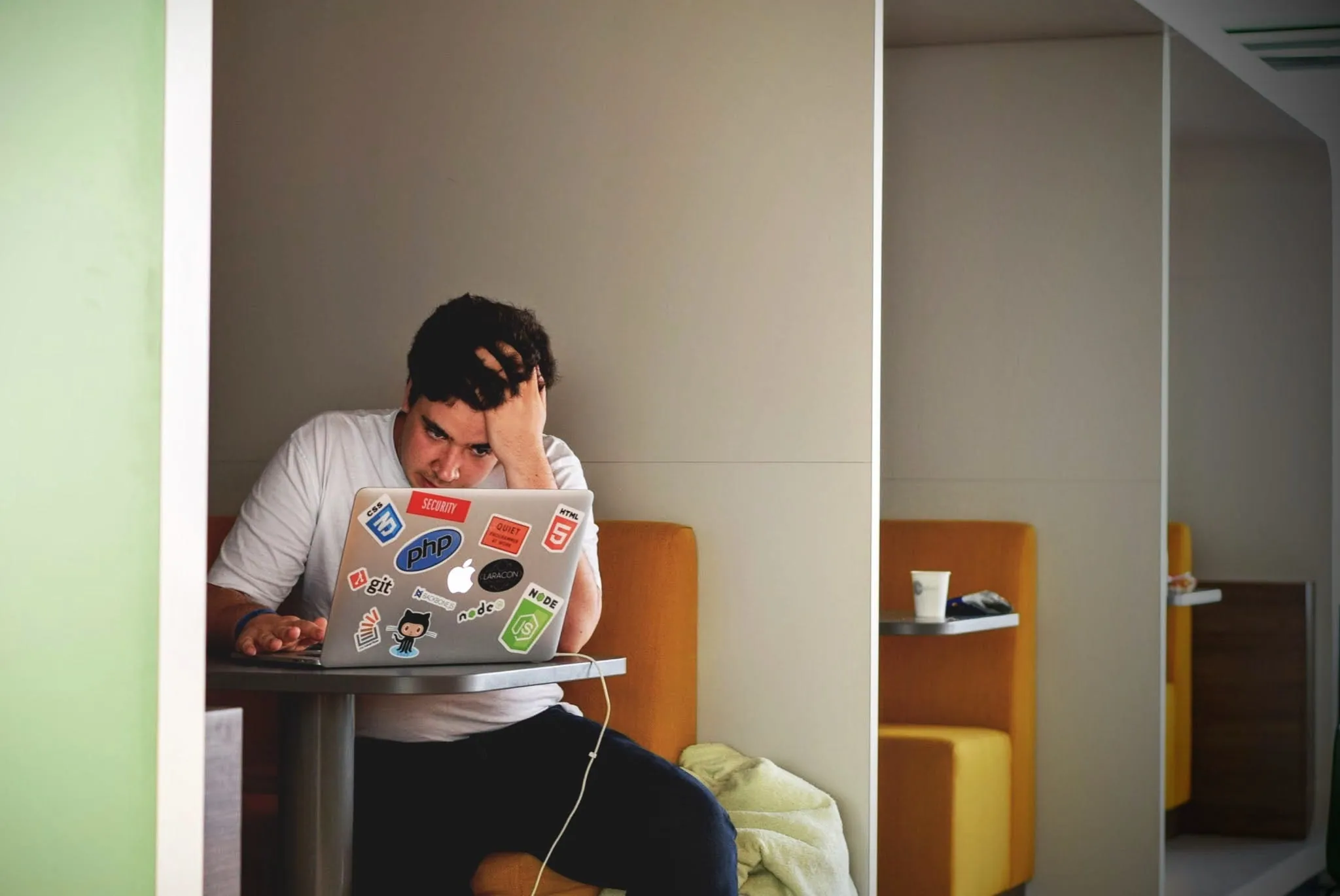 A stressed man clutching his hair while staring at a sticker-covered laptop in an office booth