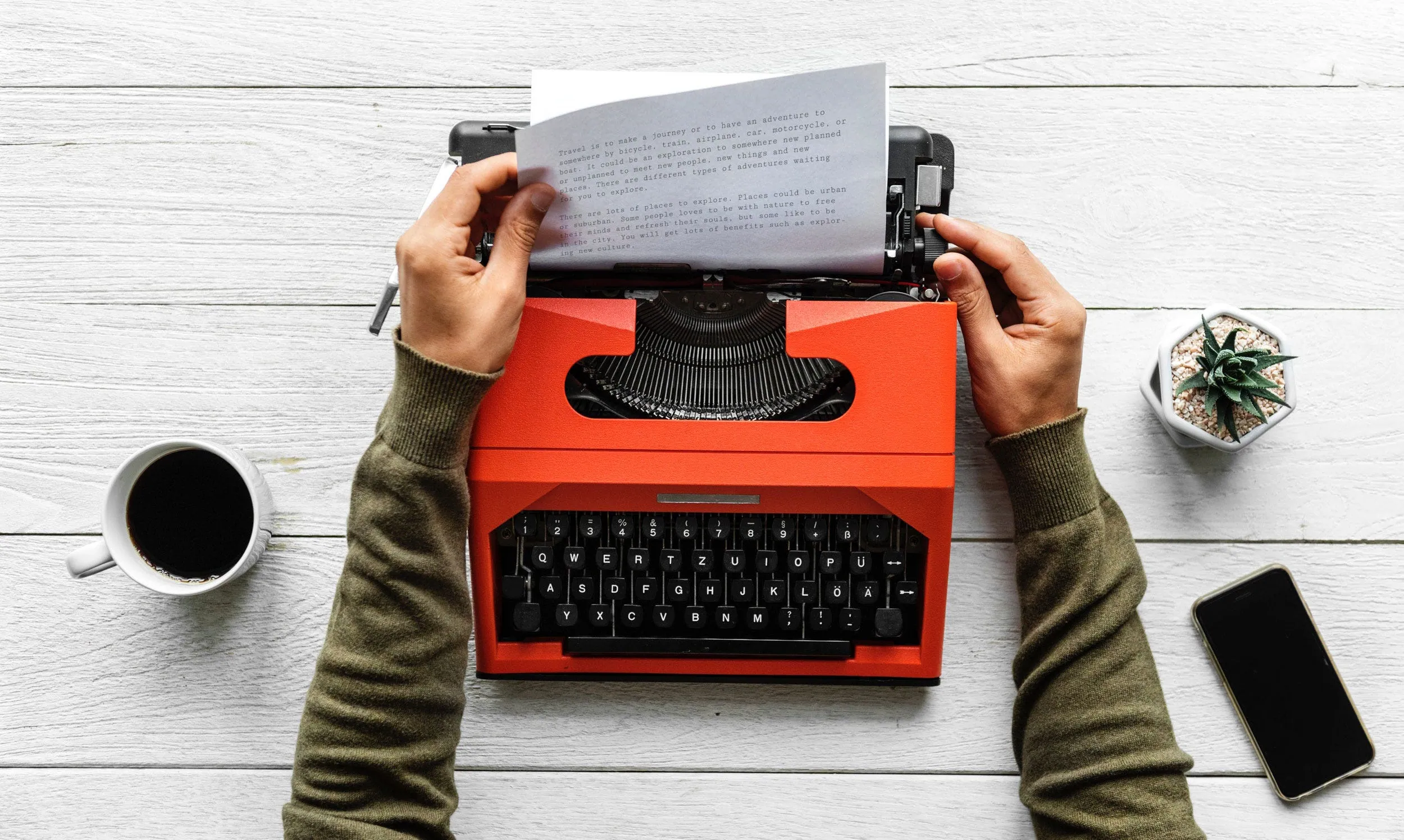 Hands typing on a red typewriter on a white desk, with a coffee cup, a plant and a phone beside it