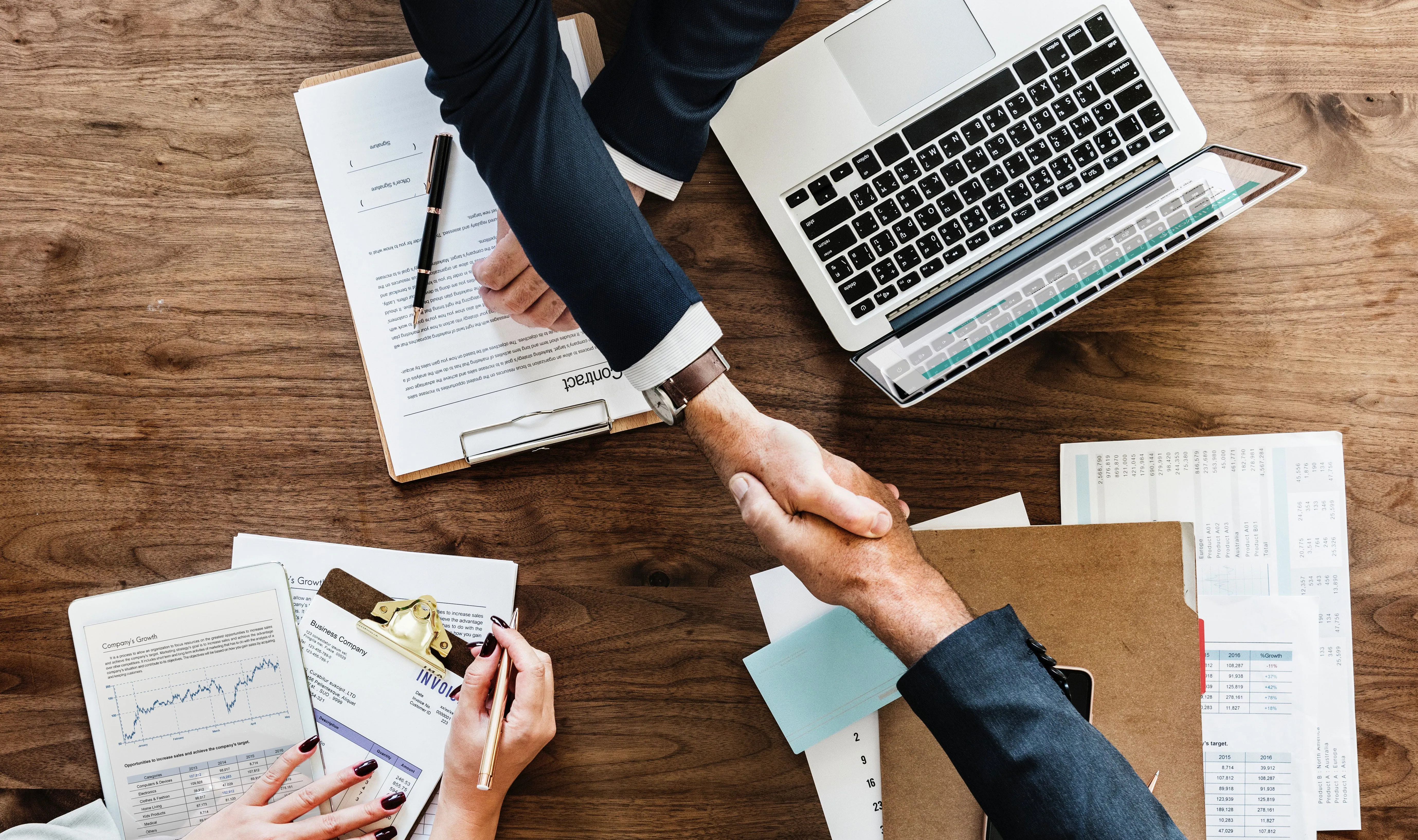 Overhead view of two people in suits shaking hands across a wooden desk covered with a contract, invoices and a laptop