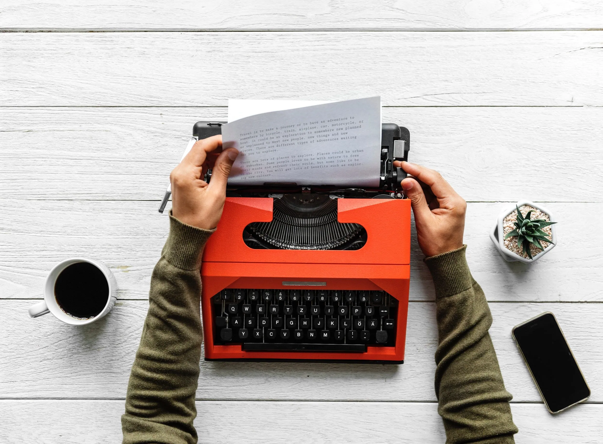 Overhead view of hands at a red typewriter with a typed page on a white wooden desk, beside coffee, a succulent and a phone
