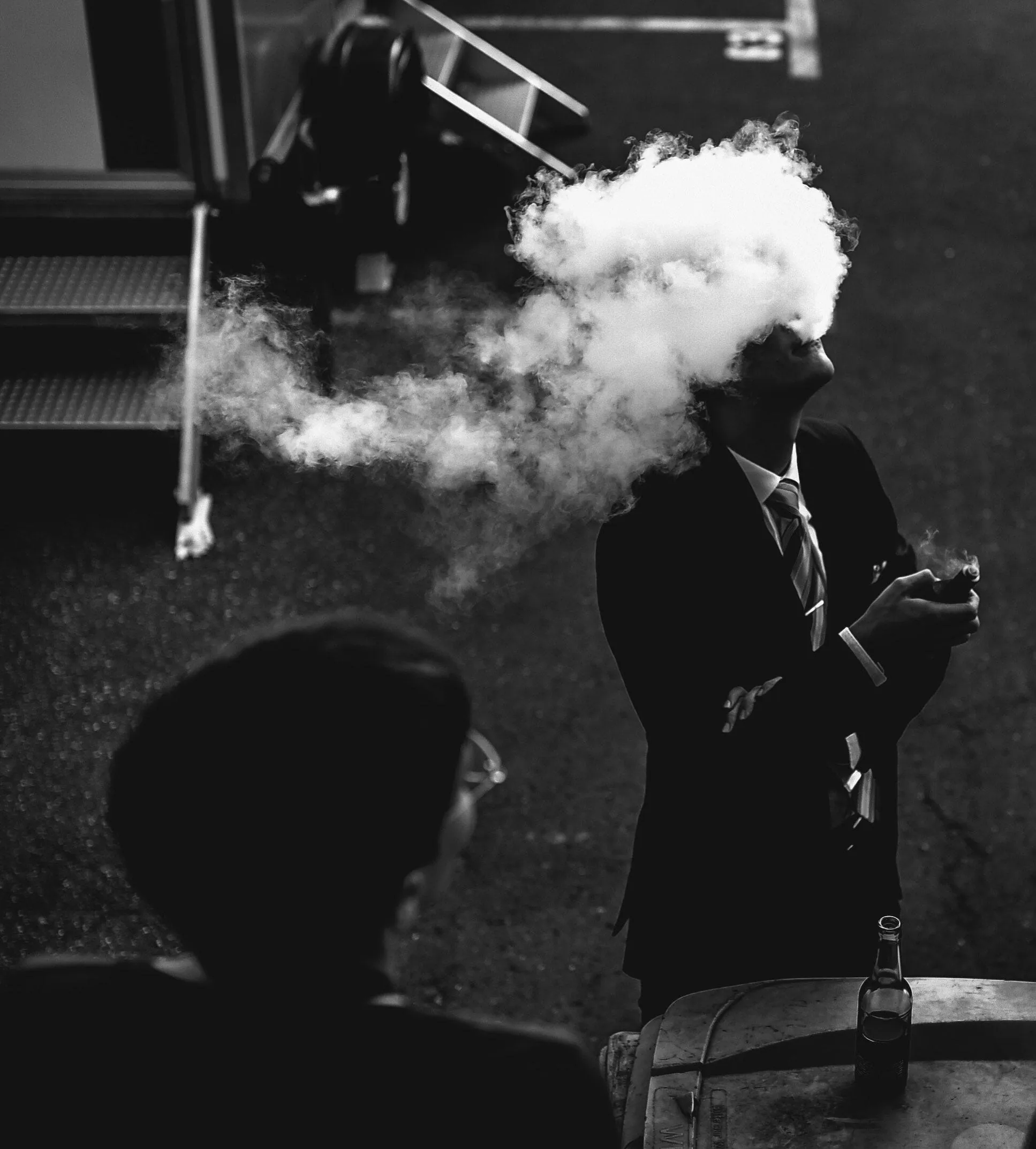 A black-and-white photo of a man in a suit exhaling a cloud of smoke