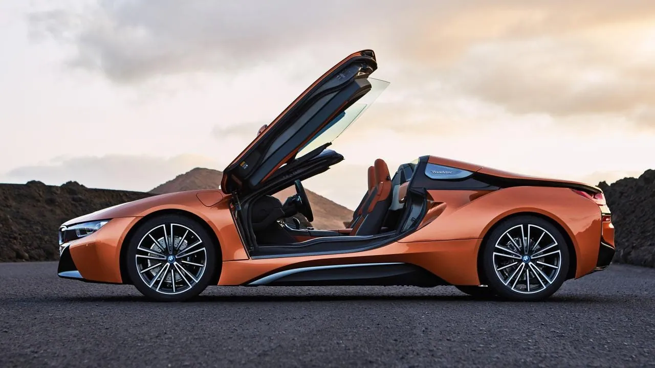An orange BMW i8 with its doors open upwards, parked on a road at sunset