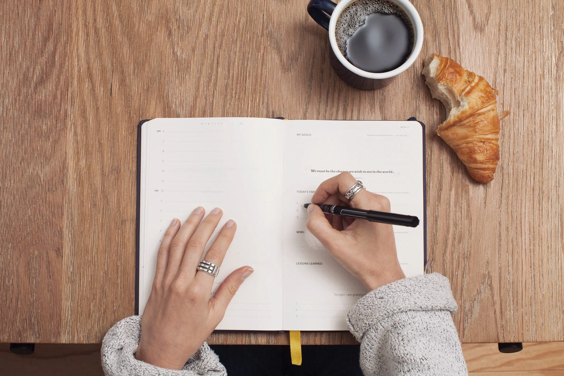 Overhead view of a hand writing goals in a daily planner beside a mug of black coffee and a half-eaten croissant