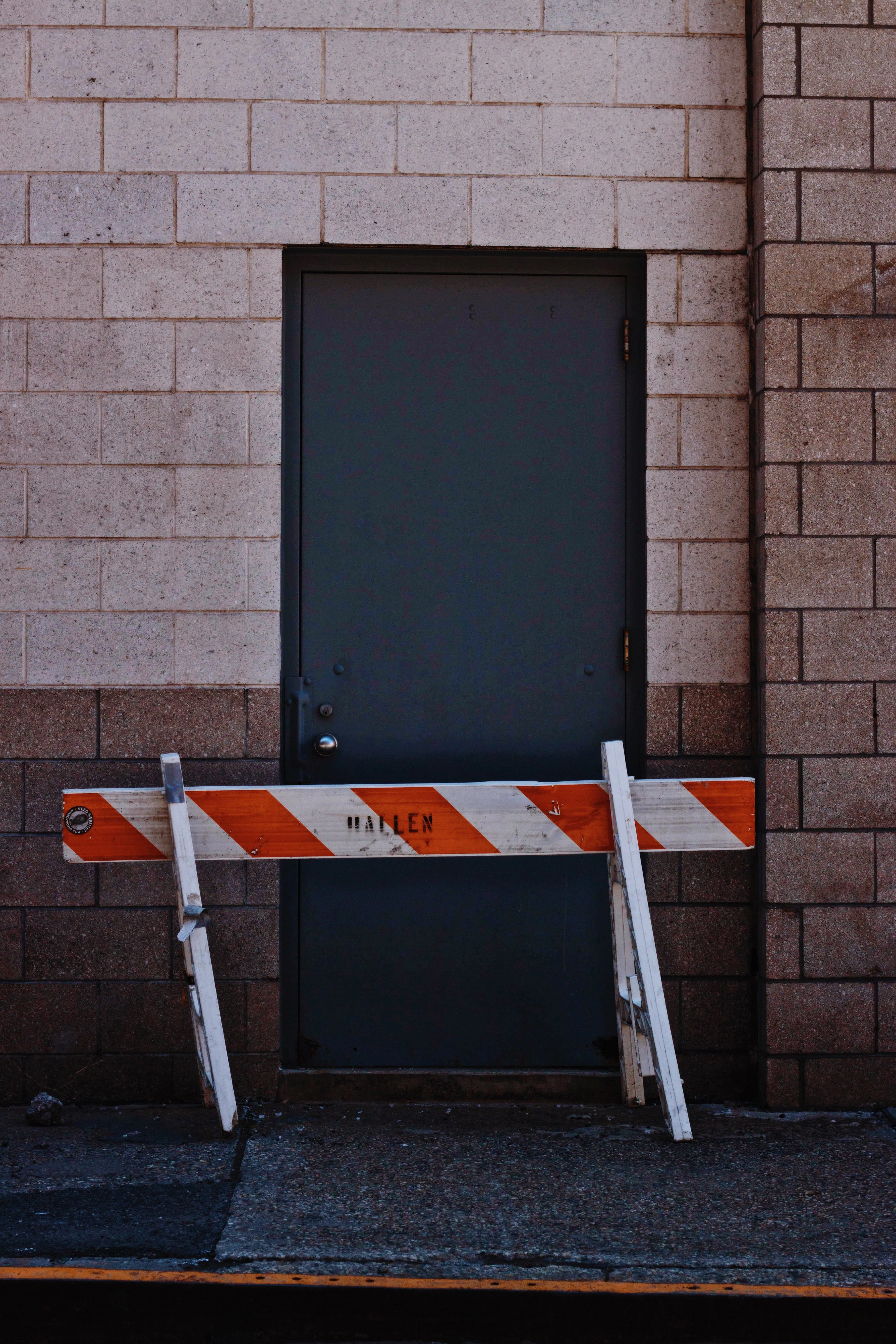 A grey door in a brick wall, blocked by an orange and white barrier