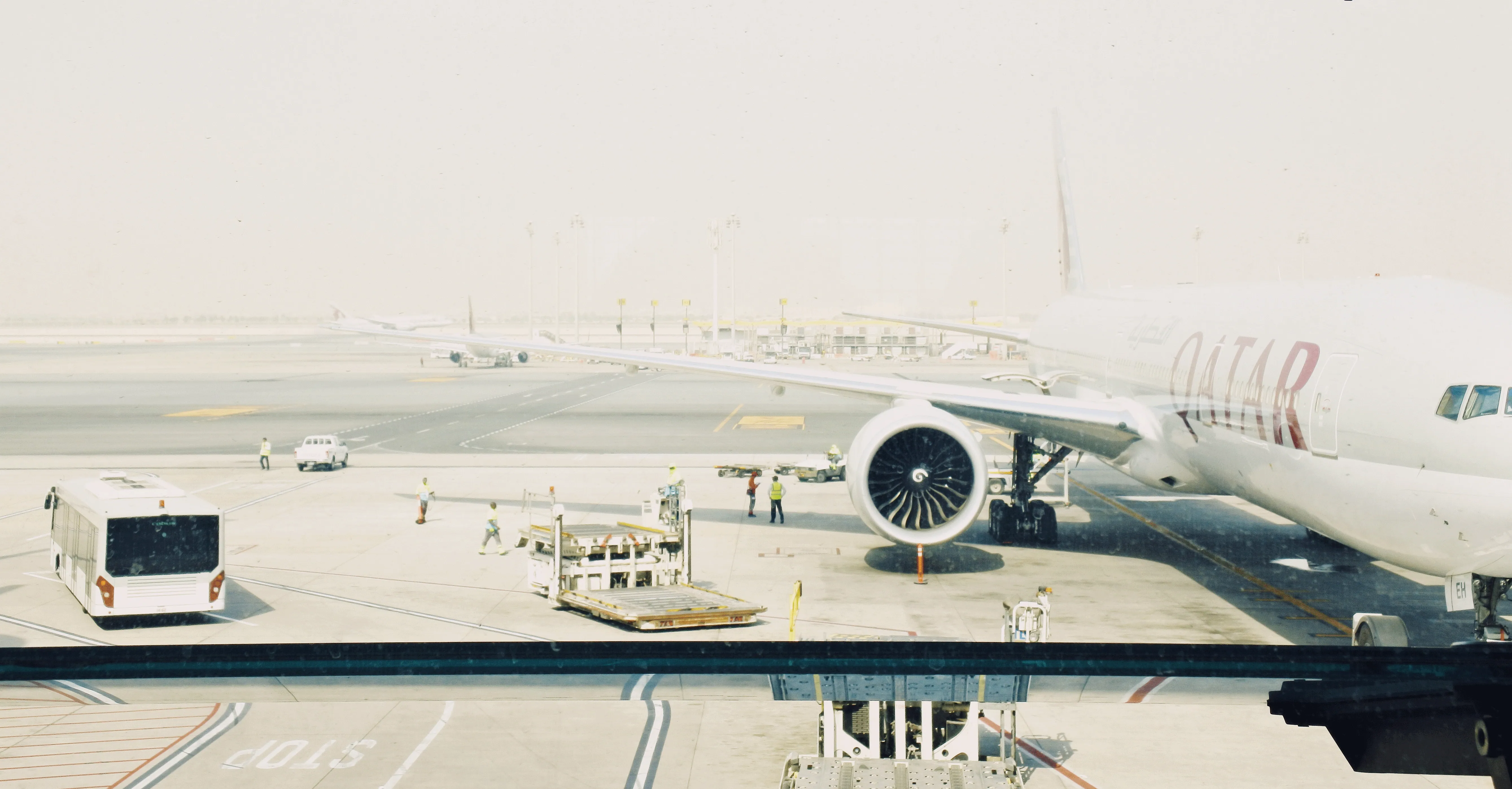 A Qatar Airways plane at the gate on an airport apron, with a bus and ground vehicles