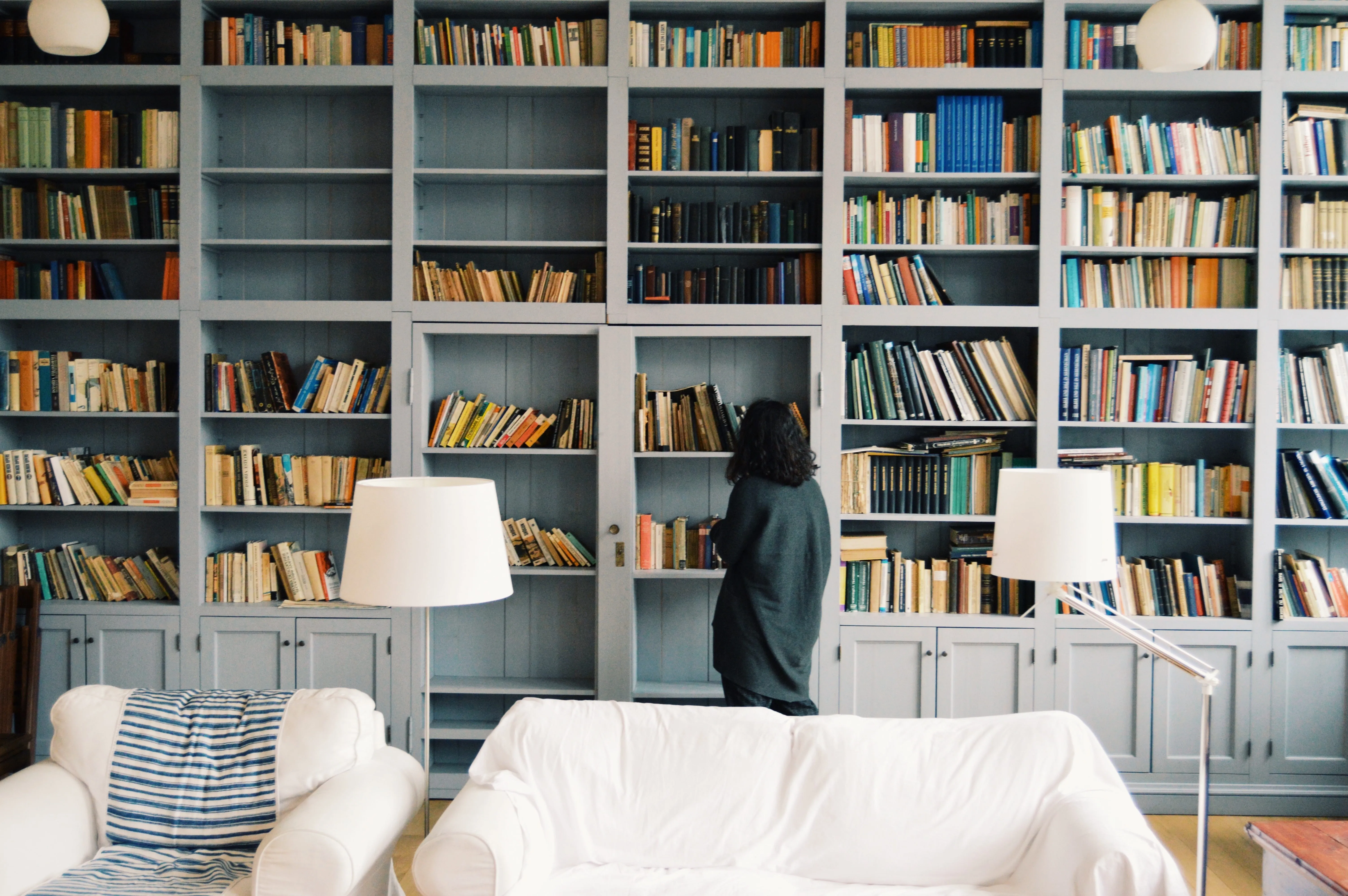 A woman browsing a wall of grey-blue bookshelves behind a white sofa and floor lamps